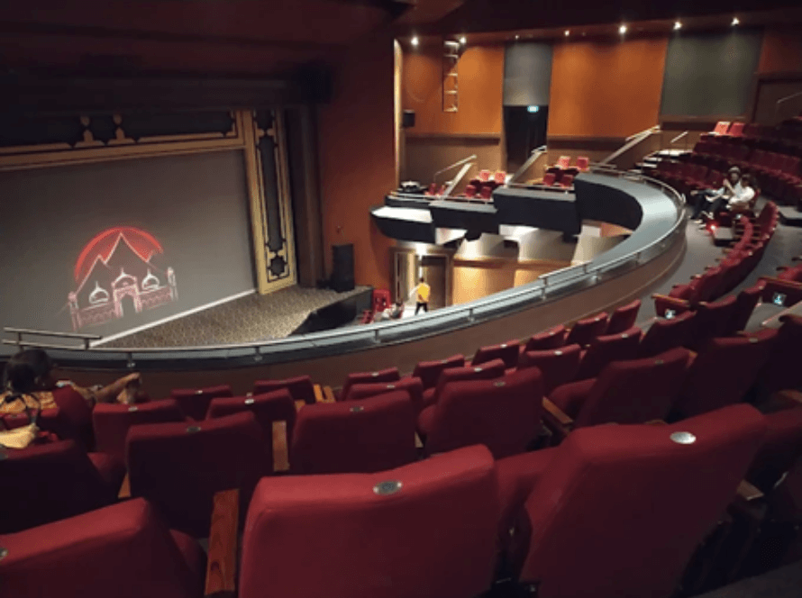 Interior view of a Balgandharva Rangmandir auditorium with red seating, curved balcony, and a stage displaying an illuminated architectural graphic